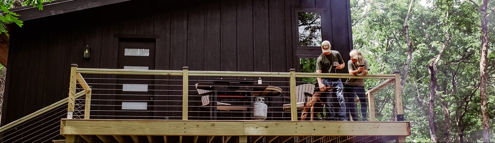 Two people on a wooden deck of a black cabin in a forested area